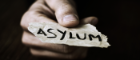 A hand holds out a scrap of paper that reads 'asylum'