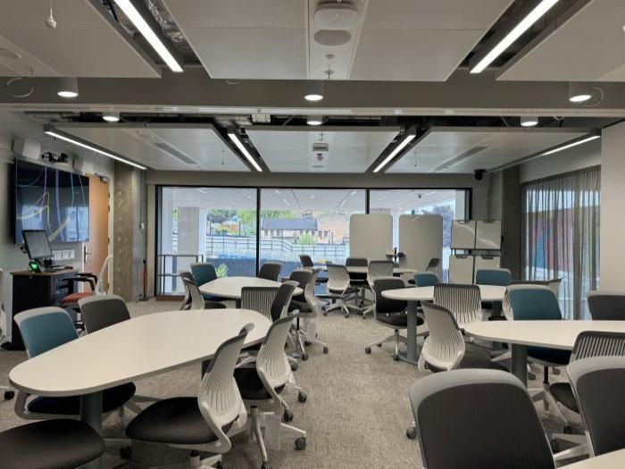 Flat floored teaching room with tables and chairs, moveable whiteboards, large video monitor, lectern, PC, and lecturer's chair.