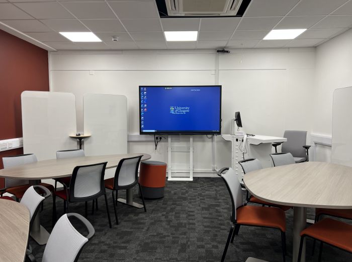 Flat floored teaching room with tables and chairs in groups, stool, video monitor, moveable whiteboards, lectern, lecturer's chair, and PC.