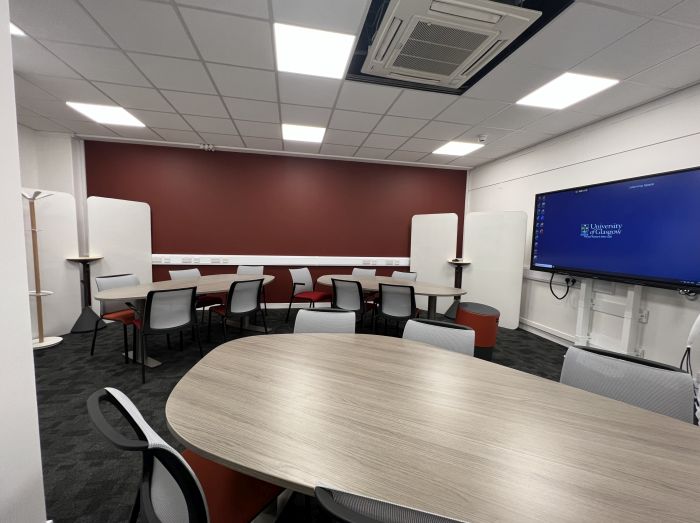 Flat floored teaching room with tables and chairs in groups, stool, video monitor, and moveable whiteboards.