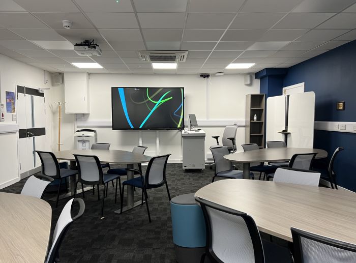 Flat floored teaching room with tables and chairs in groups, video monitor, projector, moveable whiteboards, lectern, lecturer's chair, and PC.