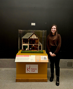 Photograph showing exhibition creator, Ruby Davidson, next to one of the exhibits. Exhibit is a 1930s dolls house. 