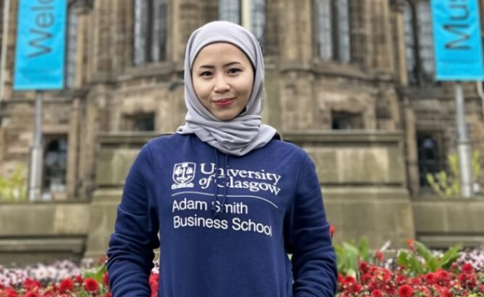 A student in an Adam Smith Business School hoodie smiles before the front face of the Gilmorehill campus