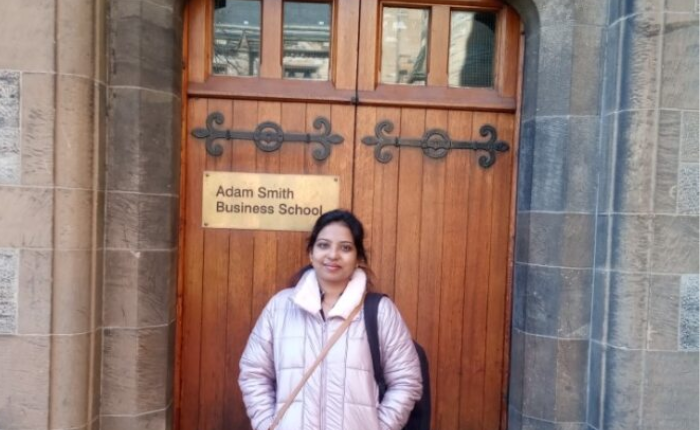 A woman in a pale pink jacket stands before an old wooden door. To her right is a gold plaque that states 'Adam Smith Business School'