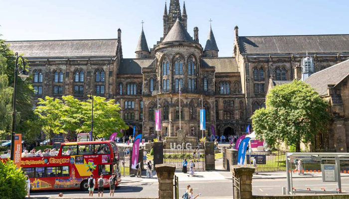 The Gilbert Scott Building on a bright sunny day, with blue and purple pop up banners out front and a tour bus passing the main gate