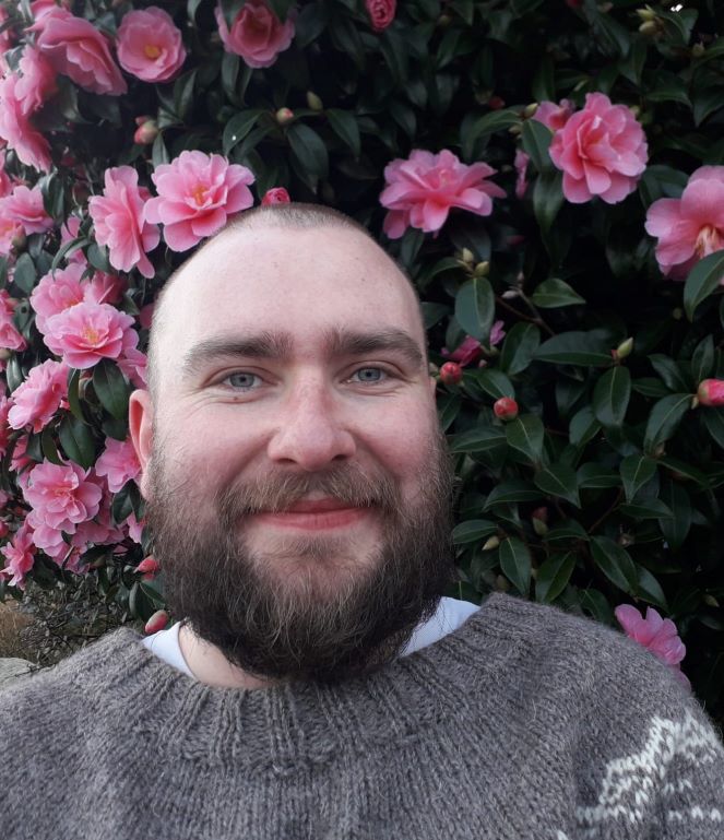 A bearded white man with a shaved head smiling in front of a pink rose bush