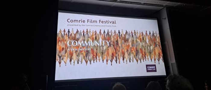Projector screen showing Comrie Film Festival poster reading 'Community: A rich tapestry' on a multicoloured background of stylised figures holding hands