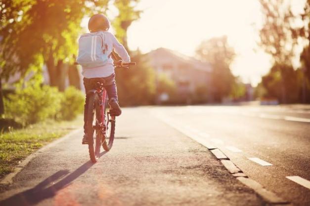 A rear view of young boy cycling along a footpath. He wears a helmet and has a backpack on.