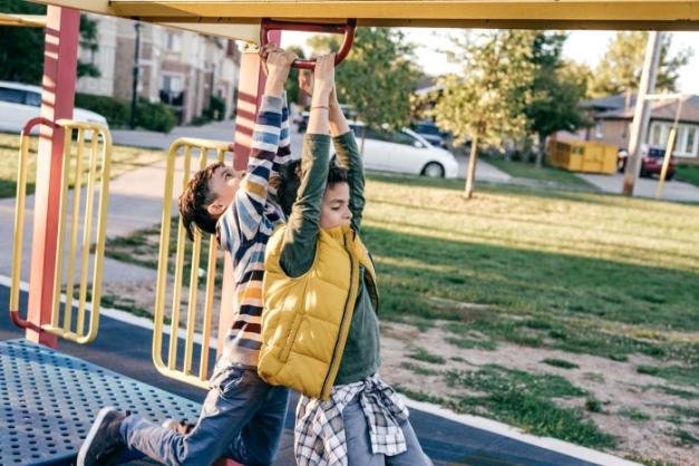 Two young boys play on a playground climber. Houses and cars are visible in the background.