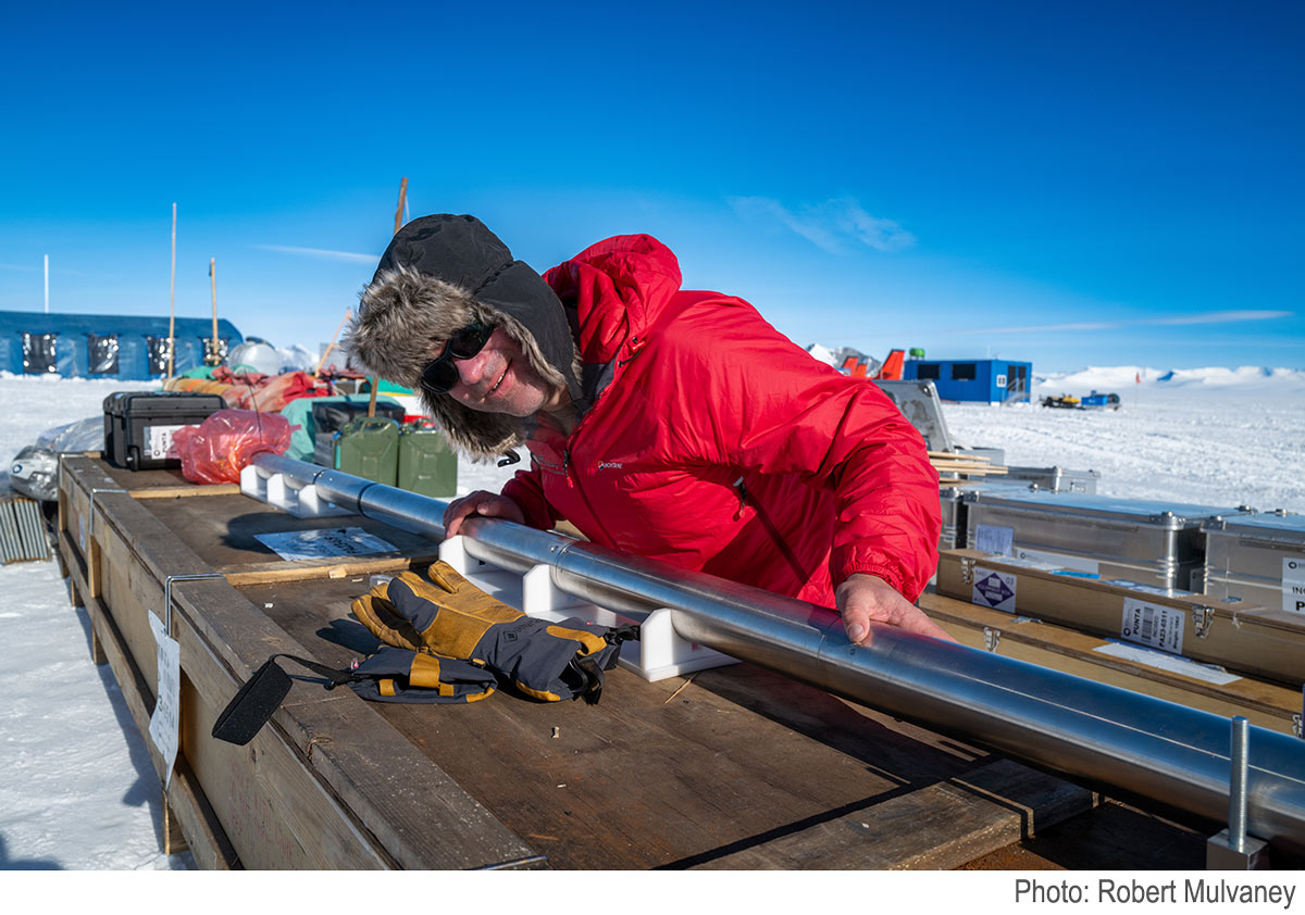 Professor Harkness picks up part of the P-RAID drill, in Antarctica, surrounded by snow and ice and under a clear blue sky.