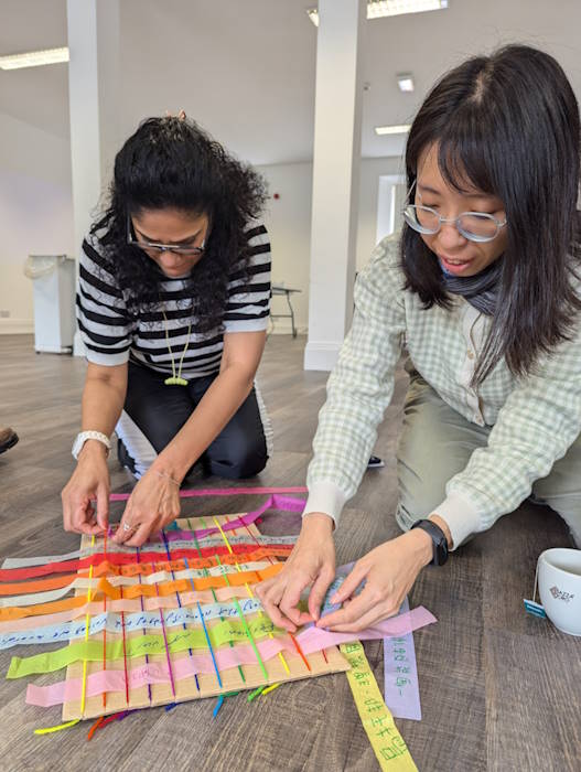 Two people crouched down weaving strips of coloured paper with words written on them