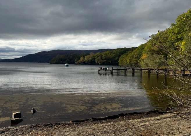 An image of people sat on a pier on the banks of Loch Lomond, taken from the share a distance away