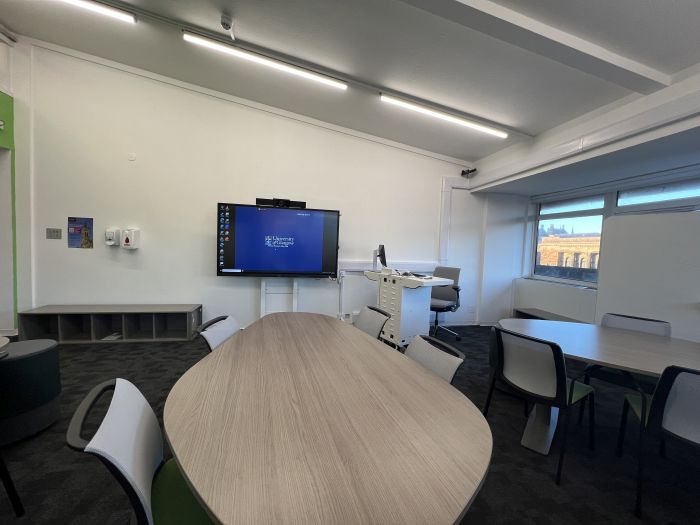 Flat floored teaching room with tables and chairs in groups, video monitor, moveable whiteboards, lectern, lecturer's chair, and PC.