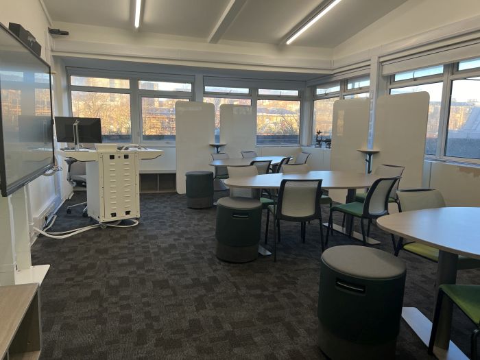 Flat floored teaching room with tables and chairs in groups, video monitor, moveable whiteboards, lectern, and PC.