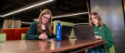 Two students studying at a table in the library. On the table is a phone, two laptops and a water bottle.