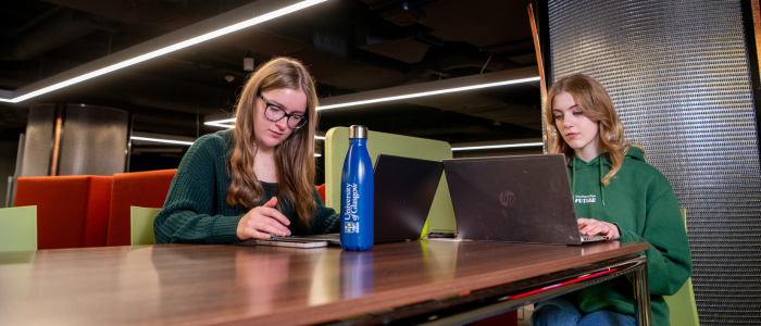 Two students studying at a table in the library. On the table is a phone, two laptops and a water bottle.