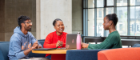 Three students sitting around a table in the James McCune Smith Learning Hub, socialising and studying. On the table is a laptop and a water bottle