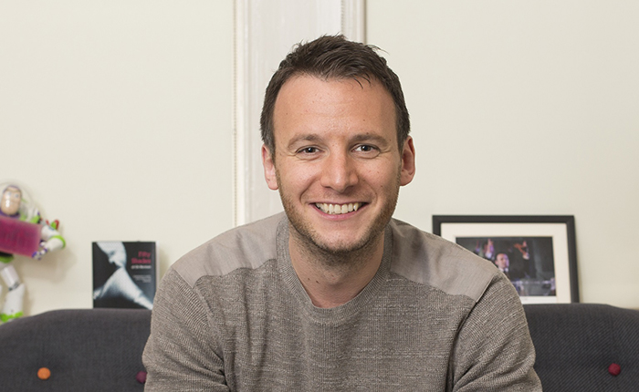 Profile photo of Oli Norman on a sofa with photo, book and an ornamental figure in the background