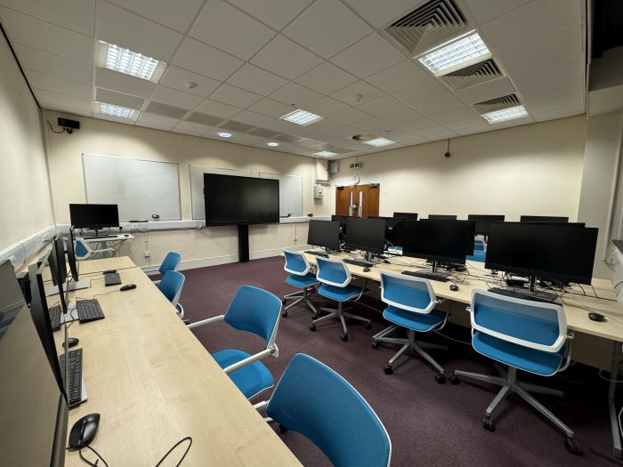 Flat floored teaching room with rows of PCs and monitors, whiteboards, large video monitor, with separate PC and chair for lecturer.