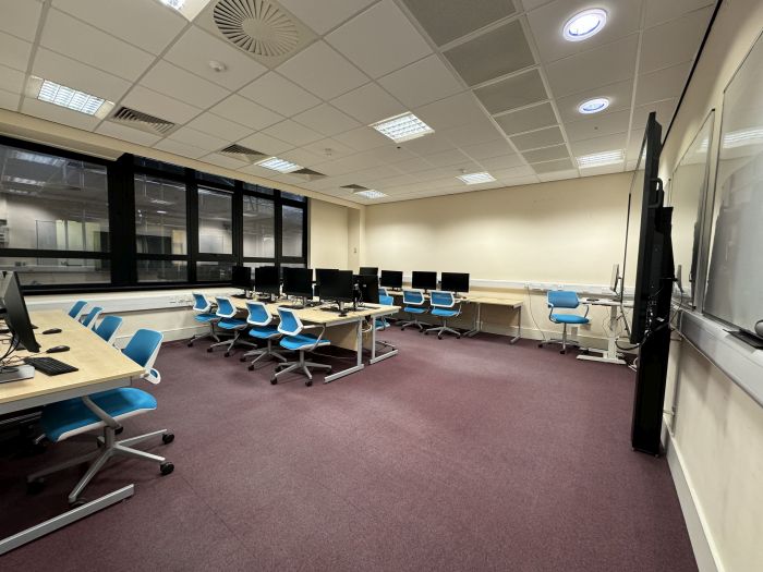 Flat floored teaching room with rows of PCs and monitors, whiteboards, large video monitor, with separate PC and chair for lecturer.