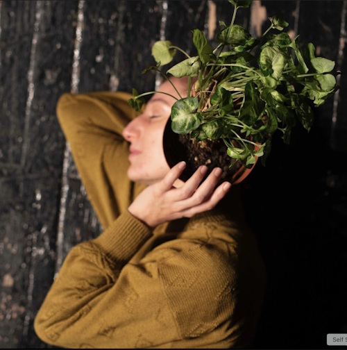 Side profile of actor Nezaket Erden holding up a potted plant at the side of her head.