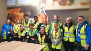 A group of thirteen University of Glasgow students and staff members wearing hi-vis vests while posing in front of a large wall covering featuring a Johnnie Walker bottle of whisky