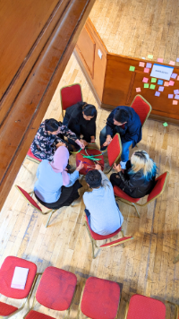 Six UofG students sitting in a circle attempting to build a phone holder out of pipe cleaners as part of a Workplace 101 session in the Glasgow University Union Debates Chamber