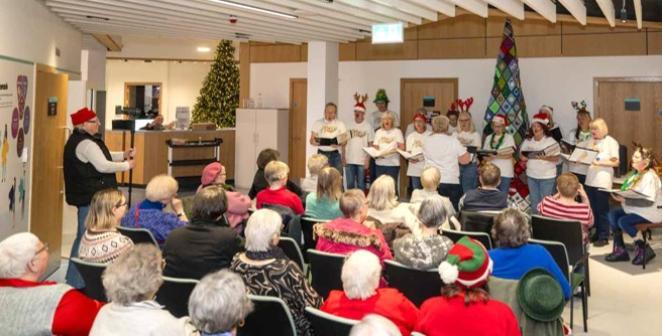 A choir are standing beside a Christmas tree singing in front of a seated audience.