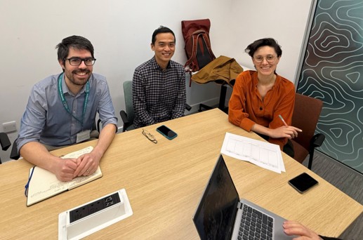 Three researchers behind a table smiling to camera. A laptop is in the foreground.