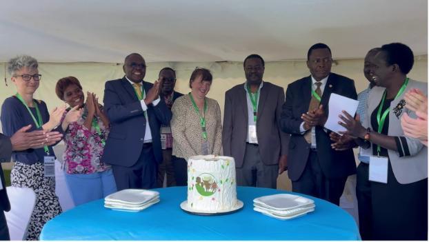 A group of formally dressed people stand around a table with a decorated white cake featuring green and brown accents. Stacks of plates are placed beside the cake, and the individuals are wearing lanyards, indicating they are attending an event or celebration.