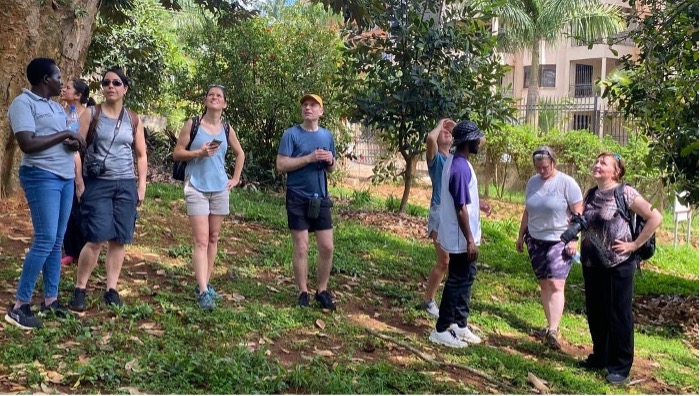 A group of seven people standing outdoors on a grassy area surrounded by trees and plants, with buildings and a fence visible in the background. They appear to be engaged in conversation or observing their surroundings.