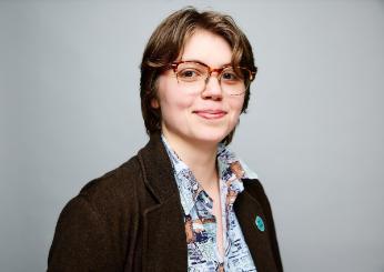 A portrait of Dr Sphinx Svensson (they/them), a person with short hair and glasses wearing a colourful shirt and brown blazer