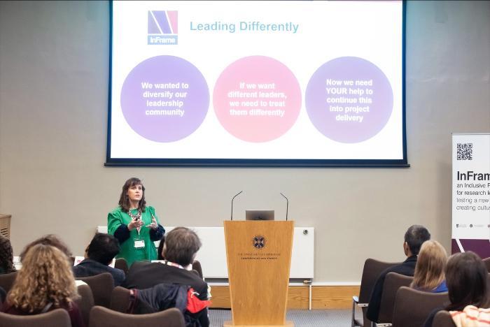 A speaker standing at a podium in a conference room presenting to an audience. Behind the speaker is a large screen displaying a slide titled ‘Leading Differently’ with three colored circles containing text: ‘We wanted to diversify our leadership community,’ ‘If we want different leaders, we need to treat them differently,’ and ‘Now we need YOUR help to continue this into project delivery.’ A banner on the right side of the room shows the InFrame logo and text.