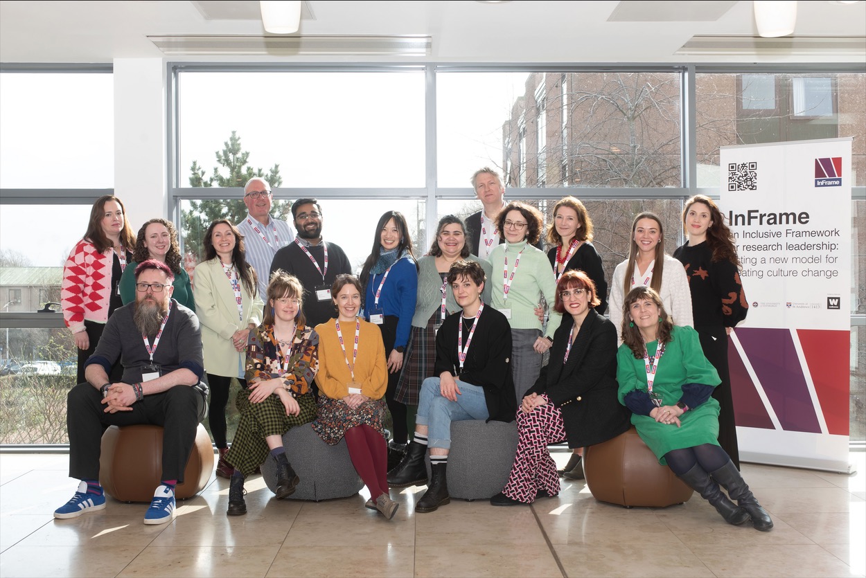 Group of people posing indoors in a bright space with large windows. They are wearing conference lanyards and a mix of colorful outfits, seated and standing in two rows. To the right, there is a tall banner displaying the InFrame logo and text about inclusive research leadership.