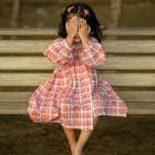 A young child sits on a wooden bench outdoors, covering their face with both hands. The child is wearing a long‑sleeved, orange and blue checkered dress, and their hair is secured with small clips. The background is softly blurred, showing the bench and ground beneath.