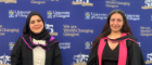 Dr Aksu and Hosay posing for a photo with graduation robes on in front of a university branded board