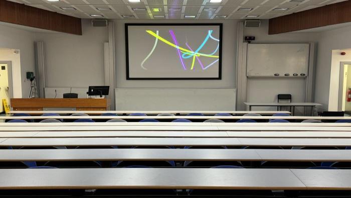 Raked lecture theatre with fixed seating, large screen, whiteboards, lectern, and PC