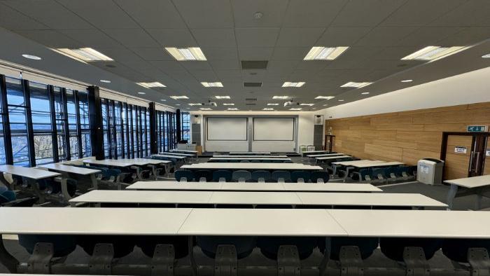 Raked lecture theatre with fixed seating, whiteboards, large screens, projectors, lectern, and PC.